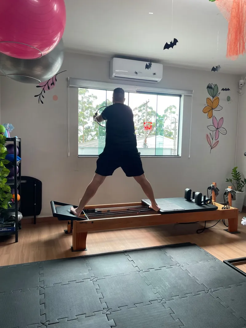 Aluno em pé no Reformer, com um pé na plataforma e o outro no carrinho, segurando o magic circle em frente à janela do studio